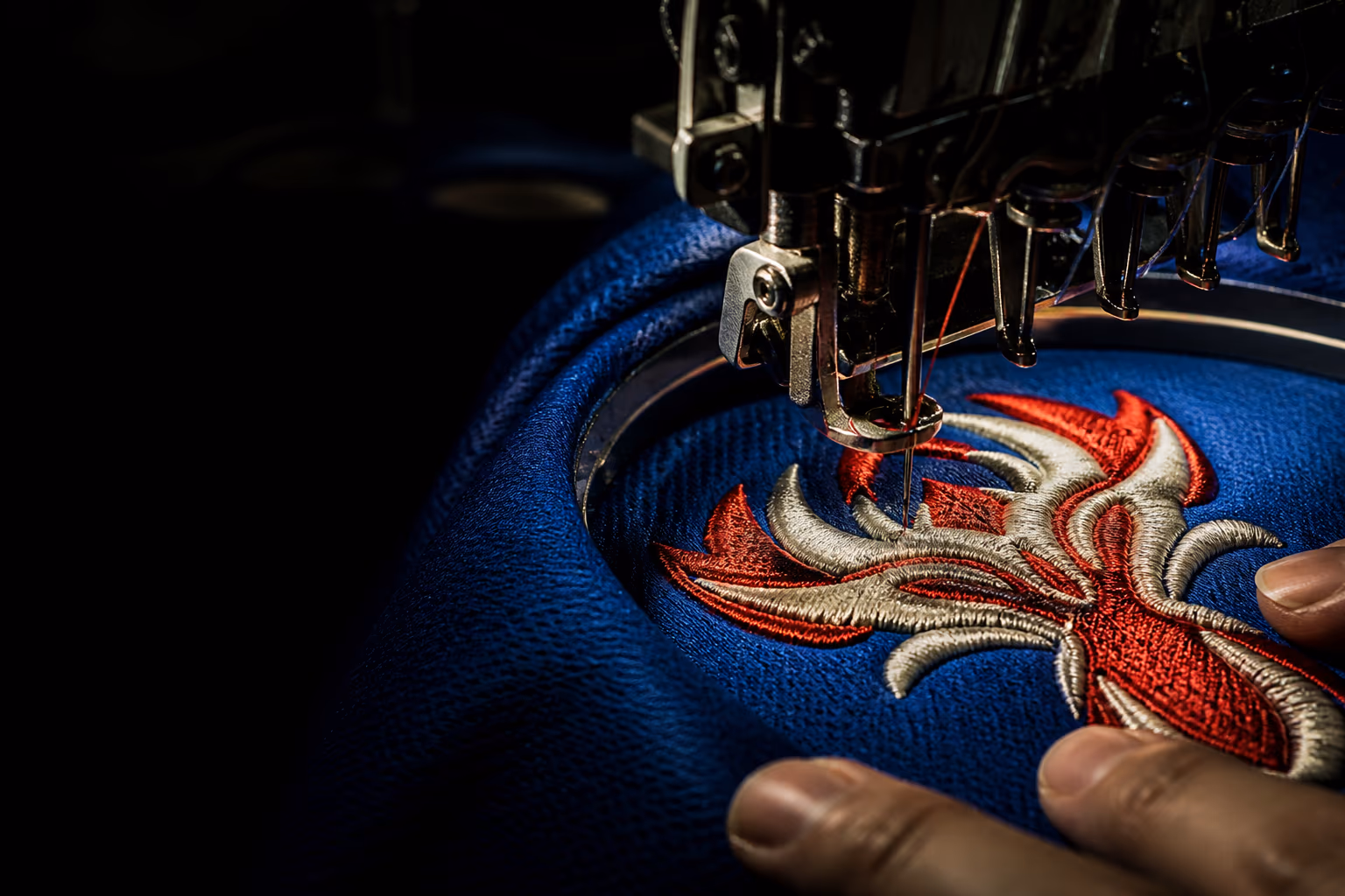 Industrial embroidery machine stitching a red and bone crest into cobalt jersey fabric